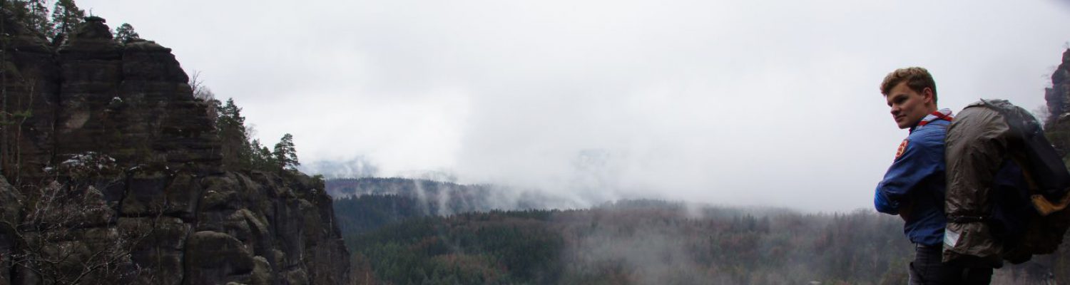 Ein nebelverhangenes Tal vor dem ein Pfadfinder steht und über seine Schulter zurück zur Kamera blickt.
